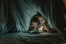 Twee kinderen lezen een boek onder een deken, bij het licht van een zaklamp